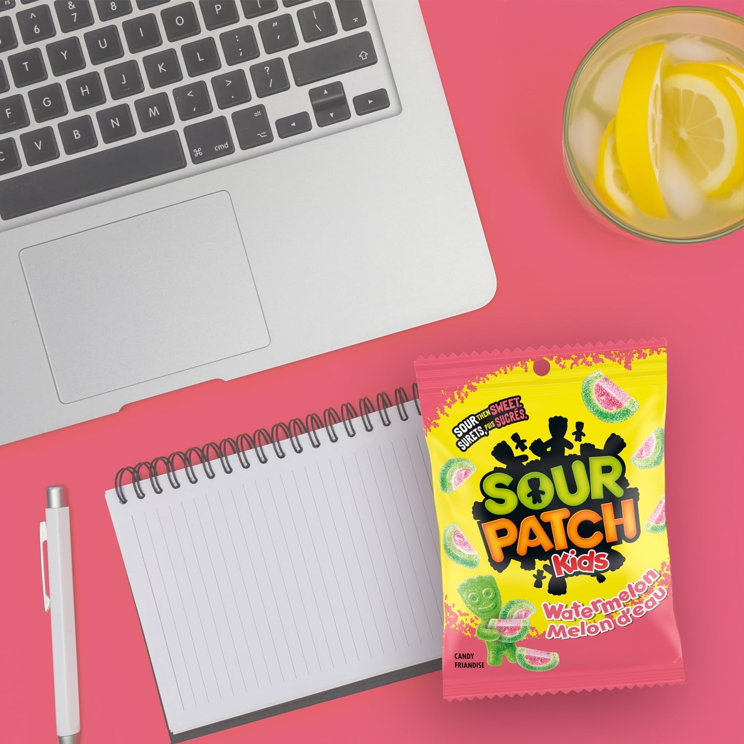 Sour Patch Kids candy package on a pink surface with a laptop, notebook, and lemonade.
