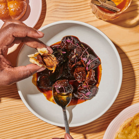 A white plate with pepperpot stew, a spoon and a hand holding a piece of pepper stew meat wrapped in flatbread on a wooden surface
