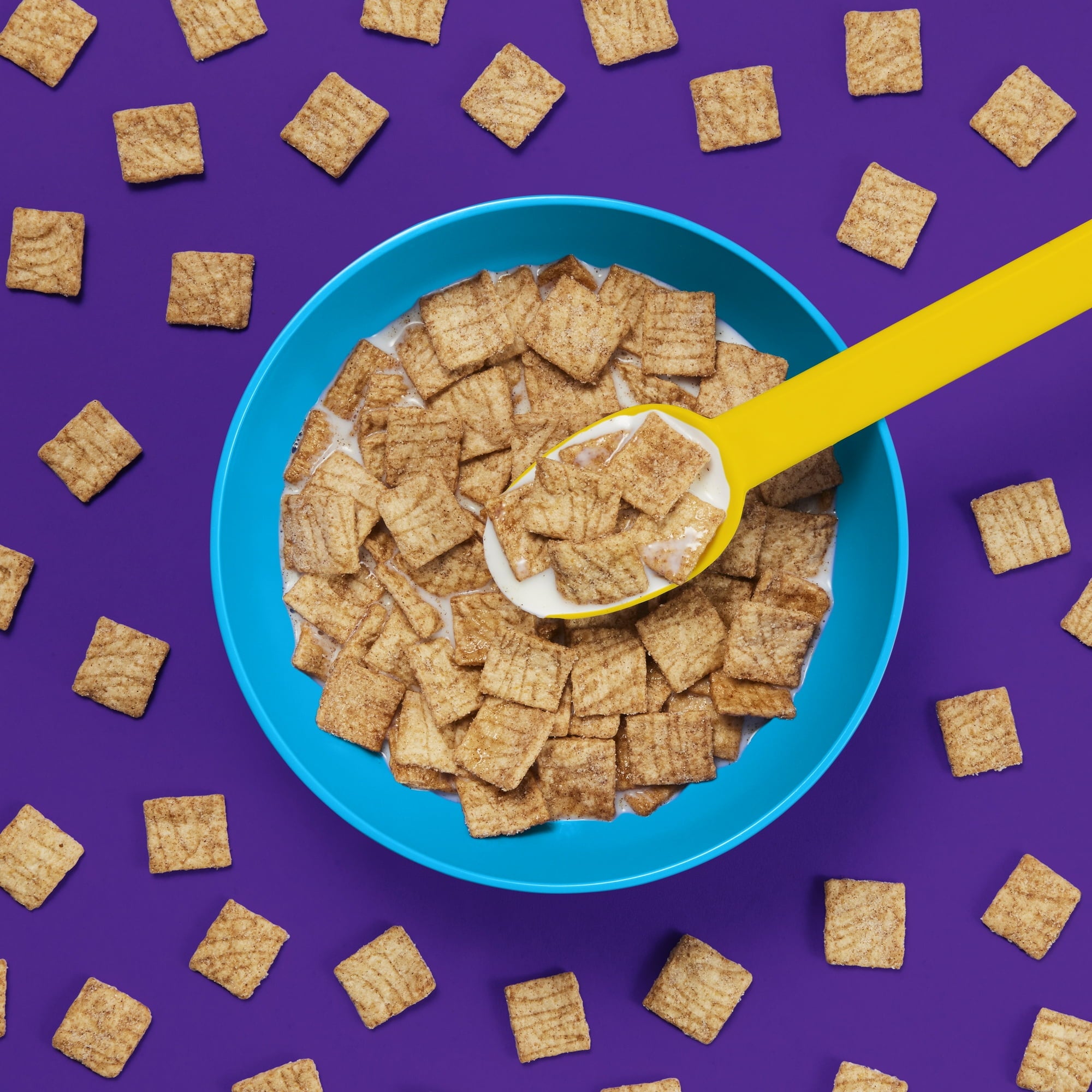 Cereal bowl with a yellow spoon on a purple background with scattered cereal pieces