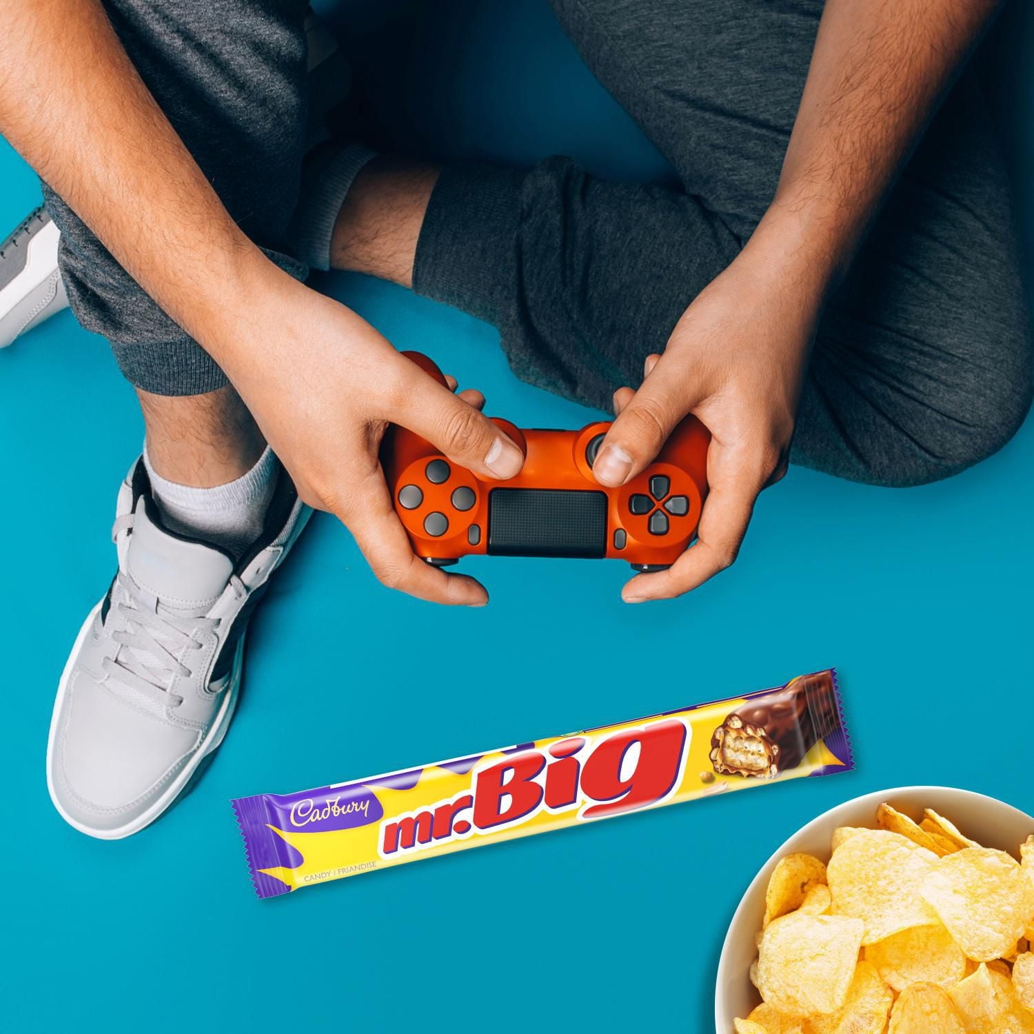 A blue surface with a mr.Big candy bar, a bowl of chips and a man holding a remote control