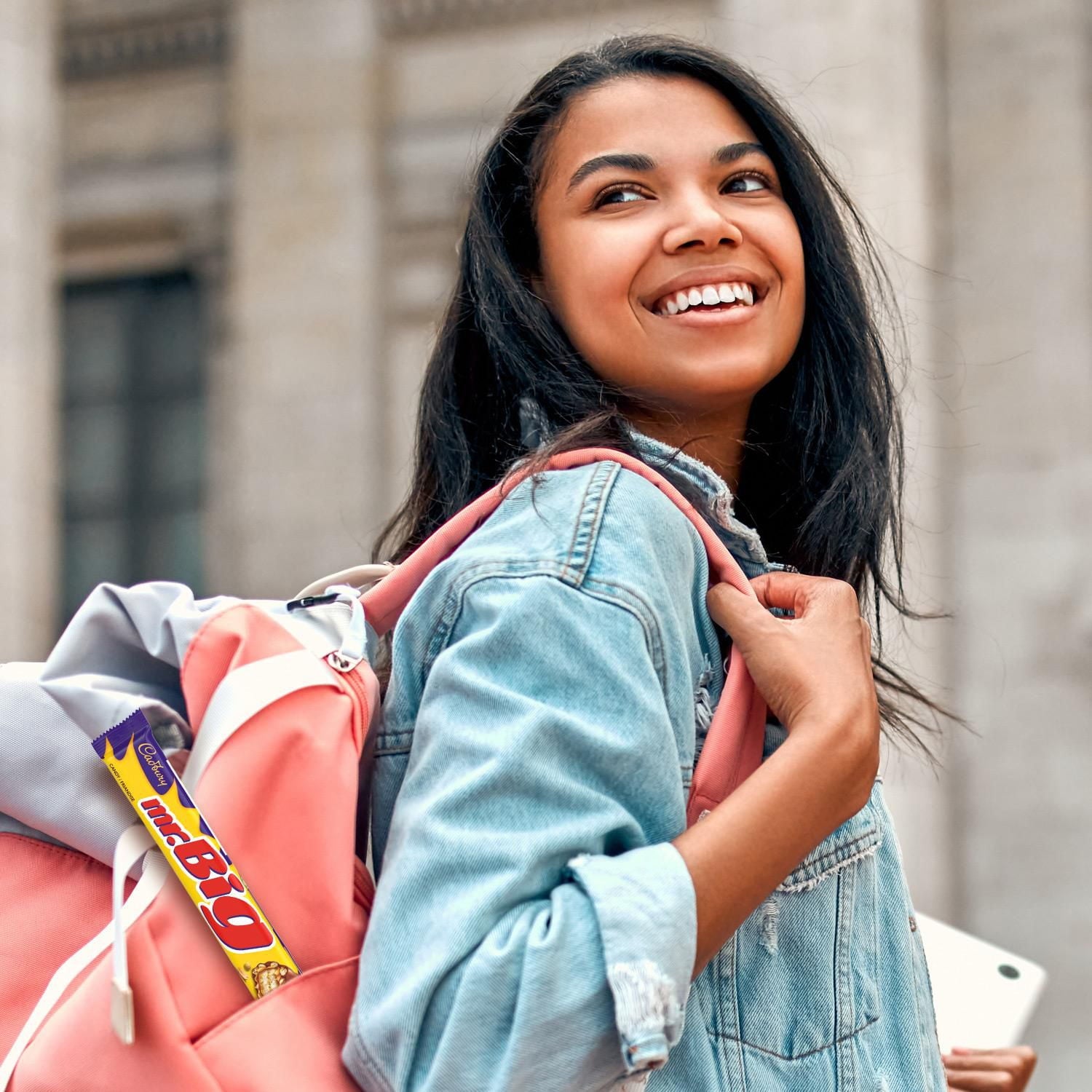 Young woman with a mr.Big candy bar in the side pocket of her pink backpack smiling outdoors