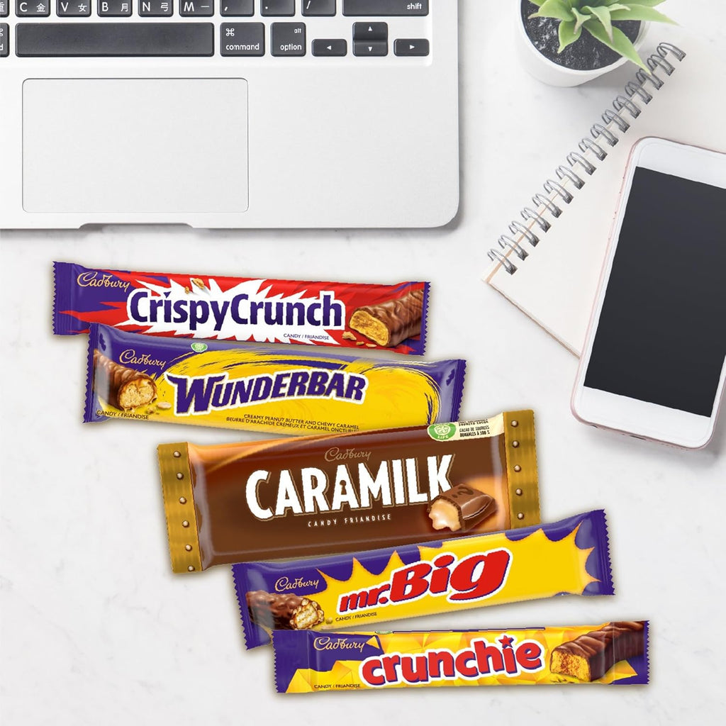 Cadbury chocolate bars displayed on a desk with a laptop, phone, and plant in the background.