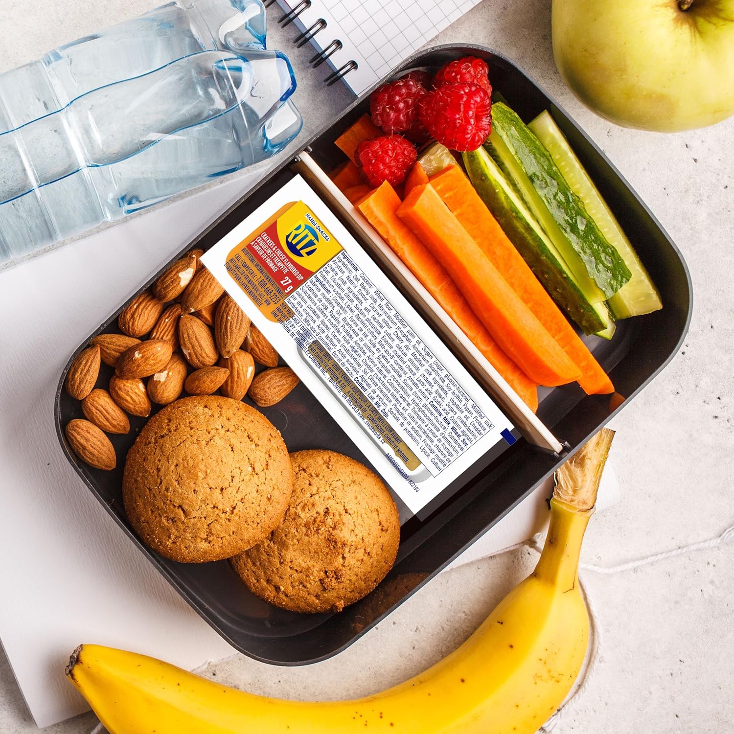 Ritz Handi-Snacks Crackers & Cheese Flavored Dip, (10x27g), 270g/9.5 oz - Lunchbox with snacks including cookies, almonds, carrots, cucumbers, and a banana on a light surface.