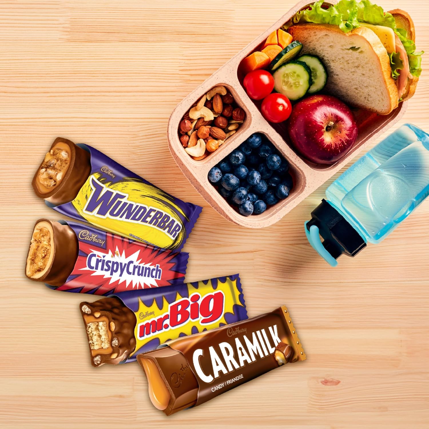Cadbury Snack-Size Bars, Wunderbar, Crispy Crunch, mr.Big and Caramilk bars beside a lunch kit filled with a sandwich, vegetables, fruit and nuts on a wooden surface
