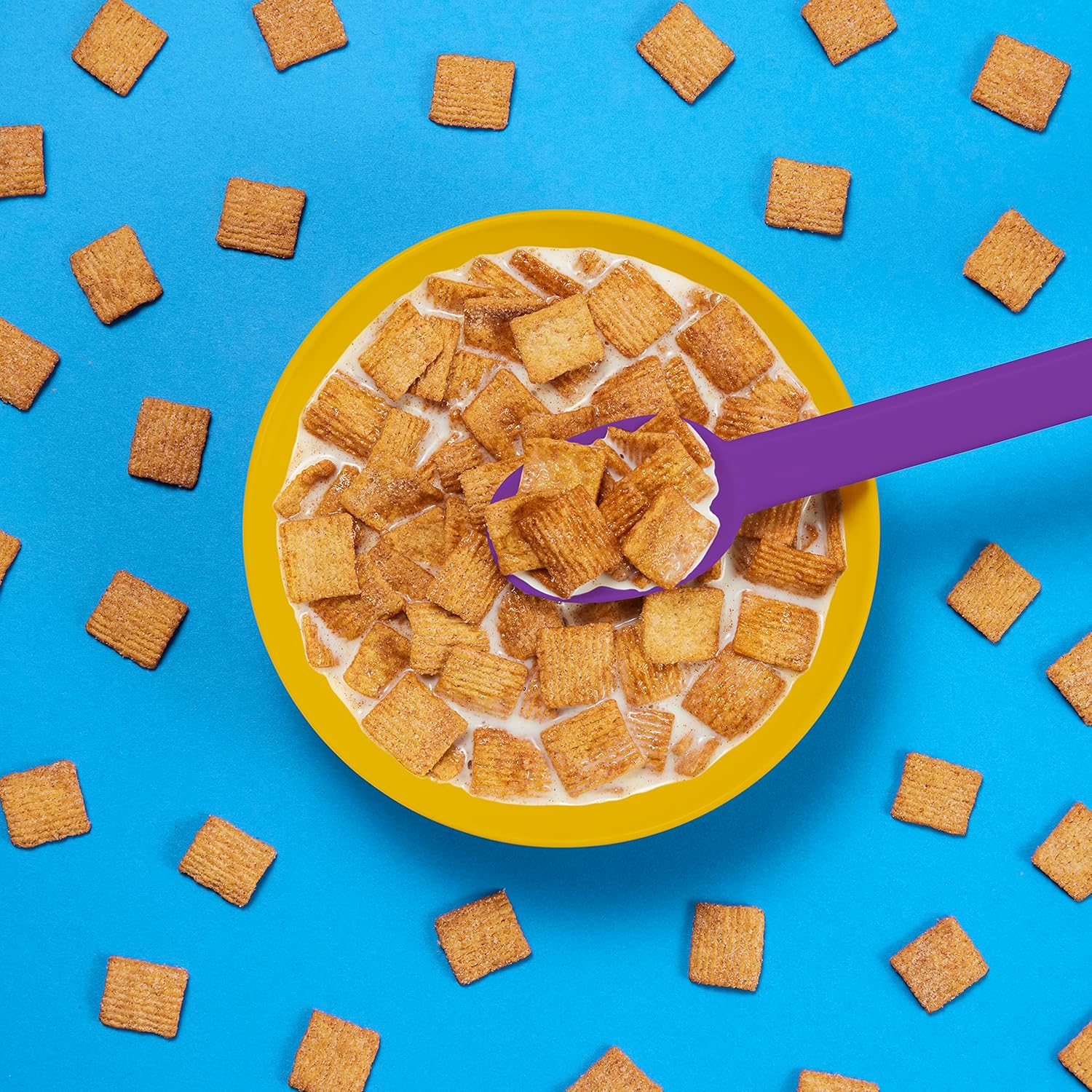 Yellow bowl of cereal with a purple spoon on a blue background with scattered cereal pieces.