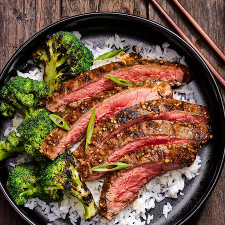 Steak with broccoli and rice on a black plate with chopsticks on a wooden table.