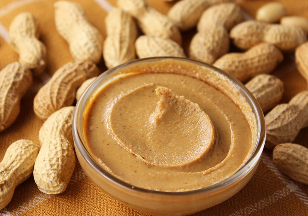 Peanut butter in a glass jar surrounded by peanuts on a wooden surface