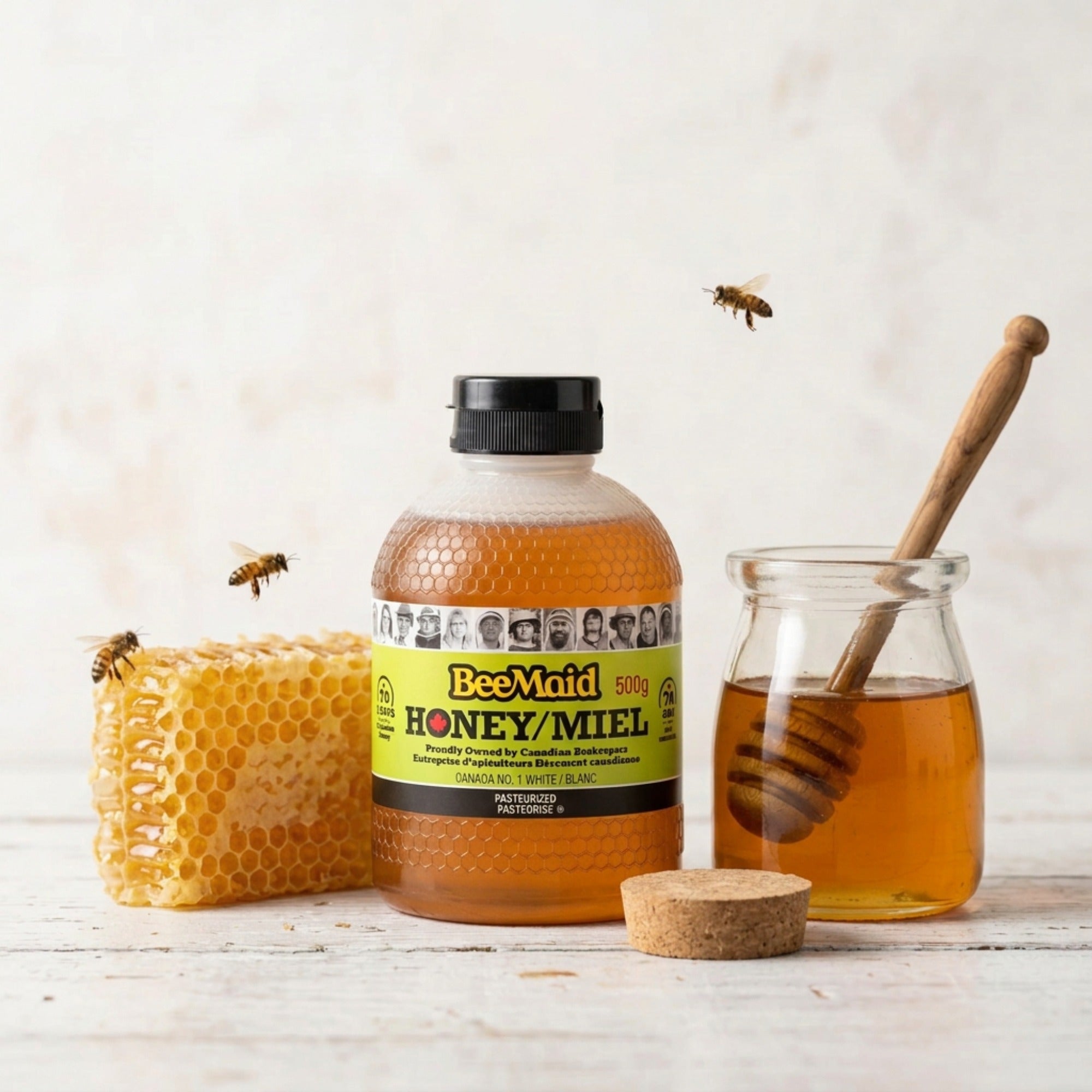 Bee Maid liquid white honey bottle beside honeycomb and a glass honey jar with a wooden dipper.