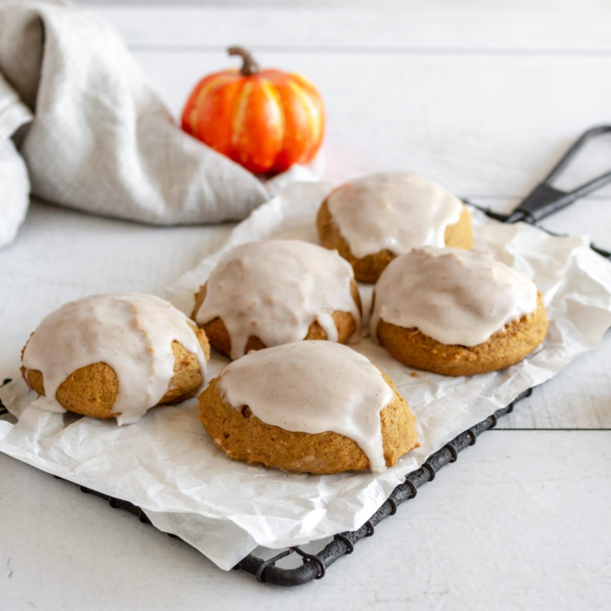 Pumpkin spice cookies topped with white glaze on a parchment-lined cooling rack, with a small pumpkin behind them.