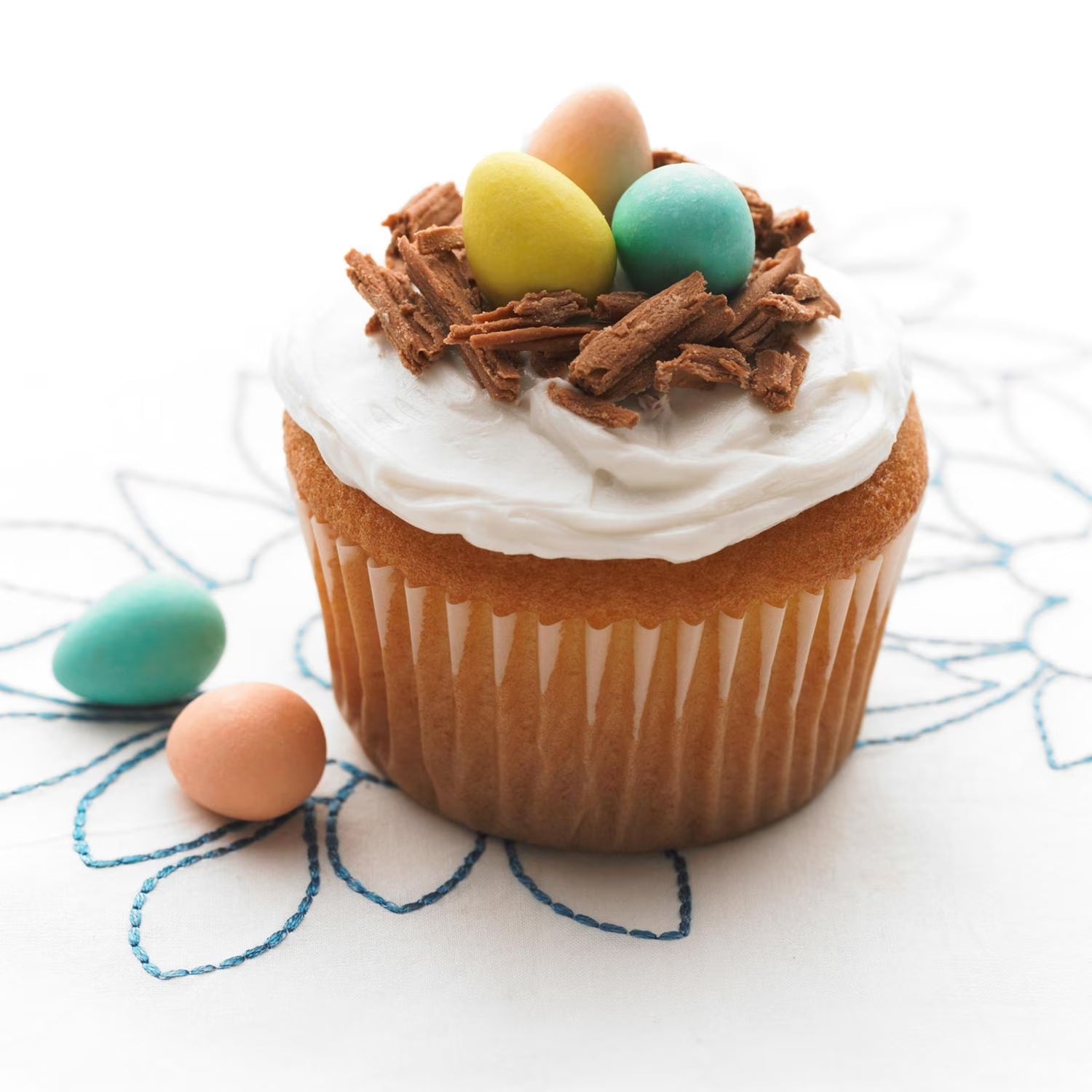 Cupcake with white frosting, chocolate shavings, and colorful eggs on a decorative white background.