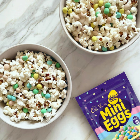 Two bowls of popcorn with Cadbury Mini Eggs on a marble surface, next to a packet of Cadbury Mini Eggs.