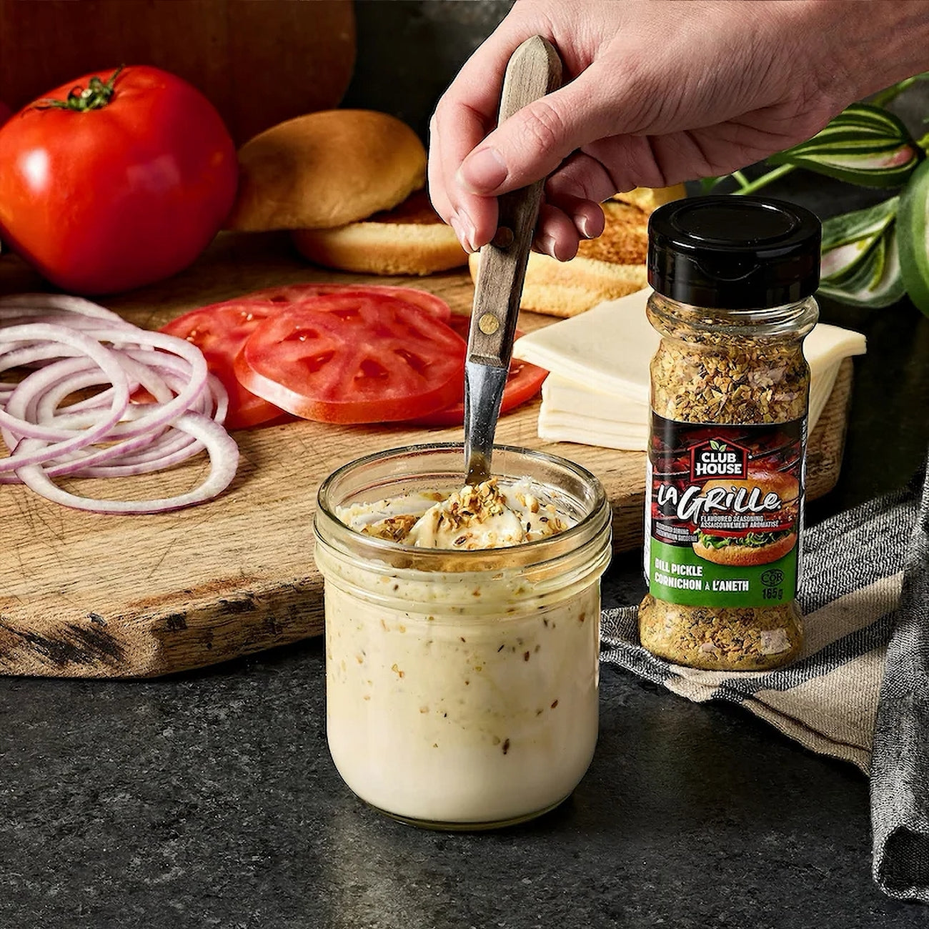 Person seasoning a jar of cream cheese with Club House La Grille Dill Pickle Seasoning on a kitchen counter.