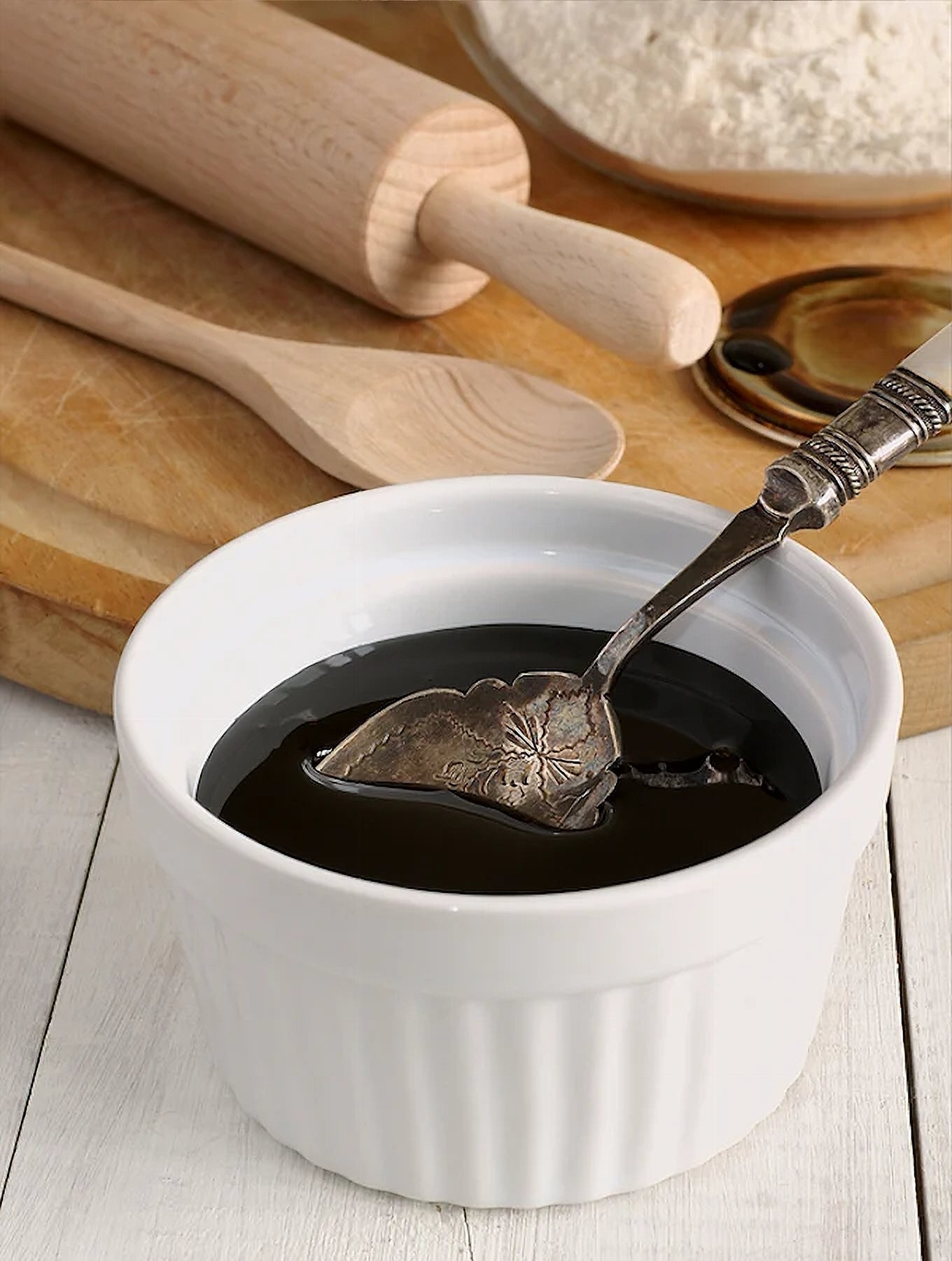 Fancy molasses in a white dish and a spoon on a wooden surface with a cooking spoon, rolling pin, and flower on a wooden board 
