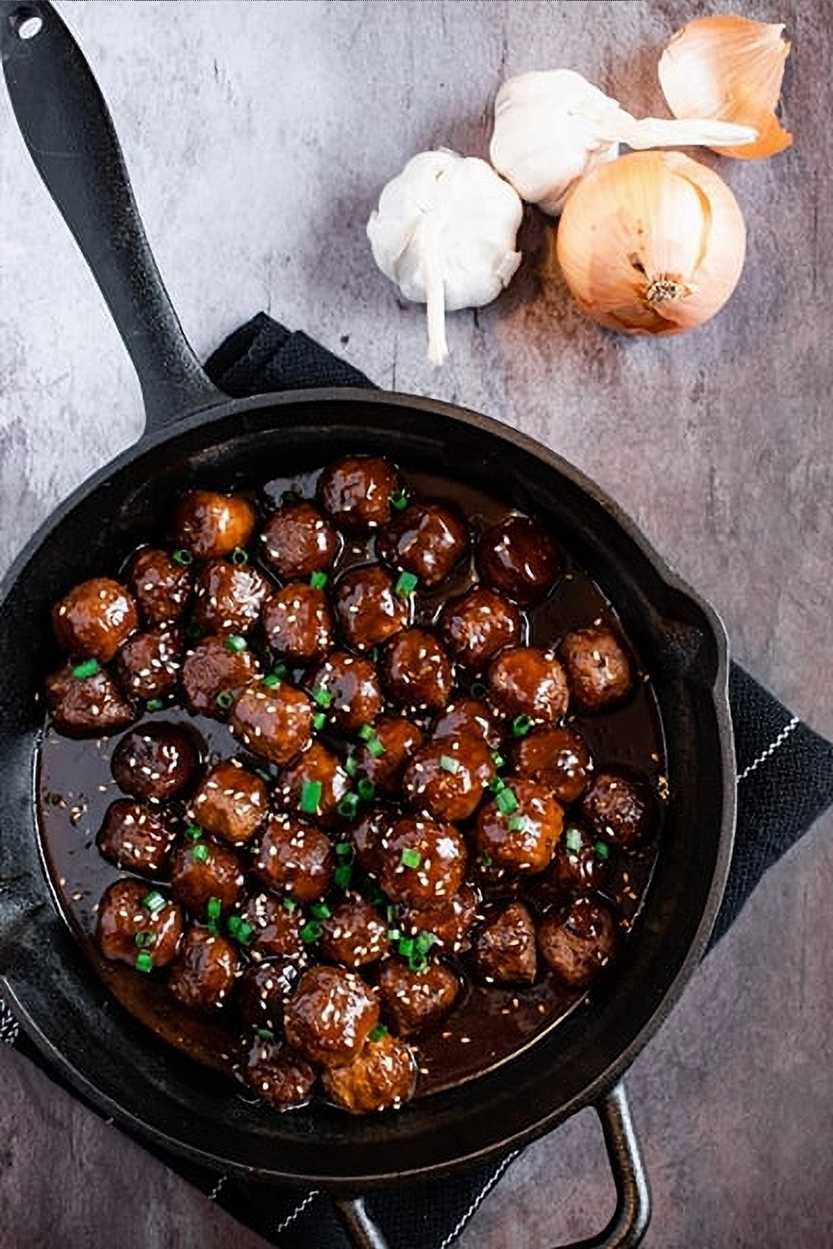 Meatballs in a molasses flavored glace in a cast iron pan on a dark towel with onions on the marble table