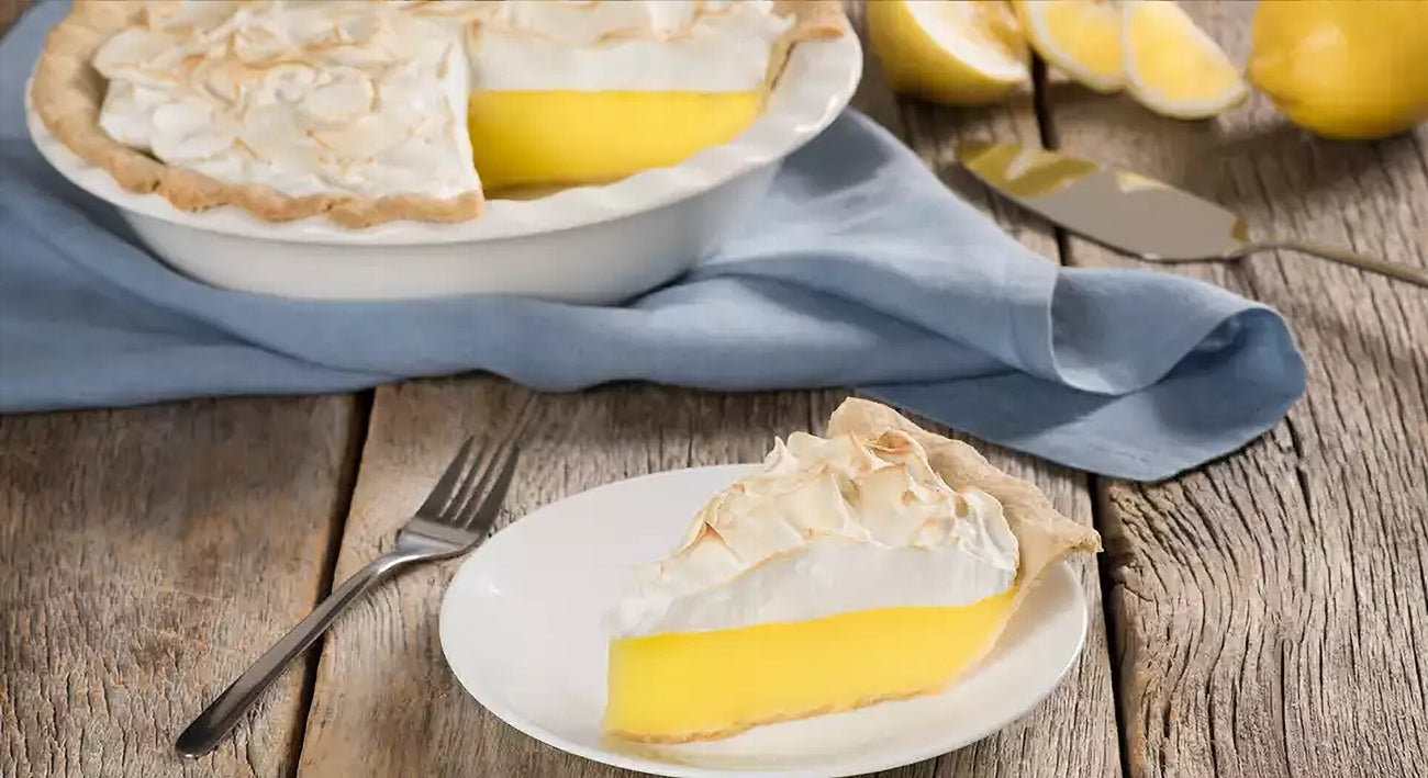 Lemon meringue pie with a slice on a plate, surrounded by lemons and a fork on a wooden table.