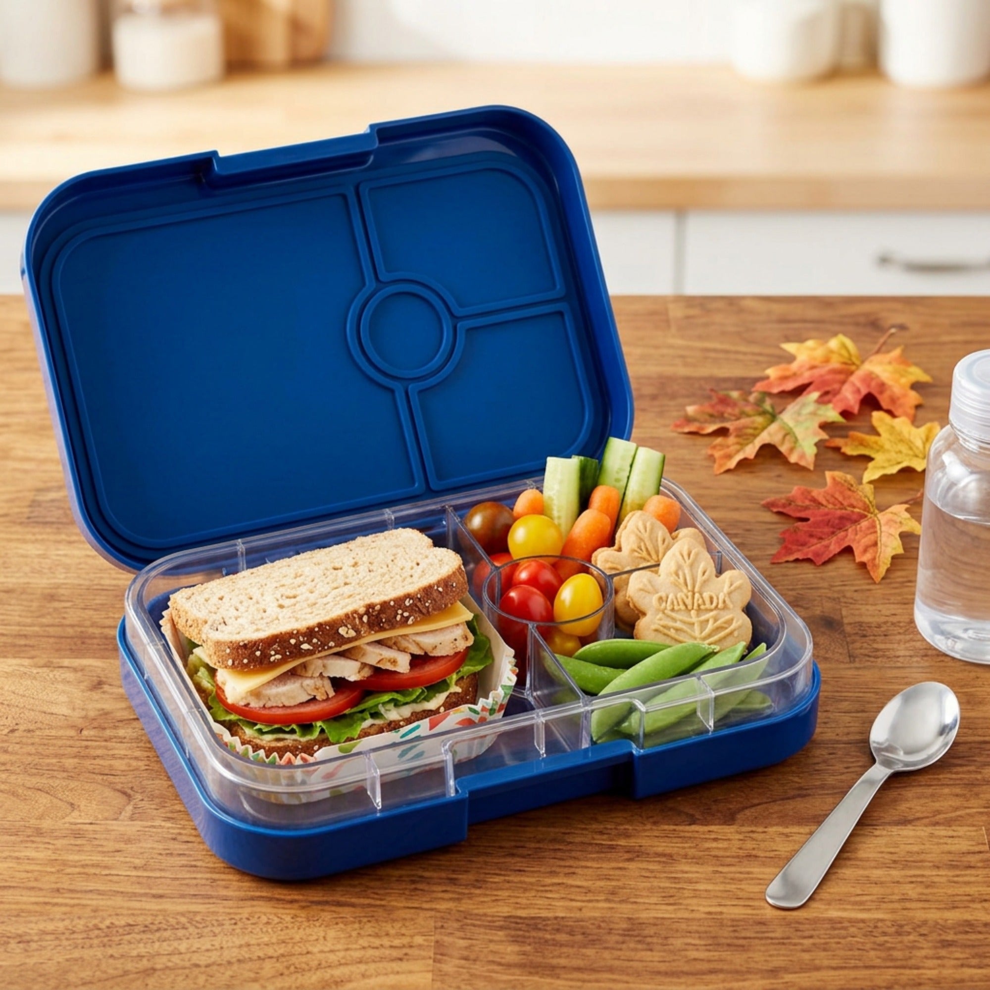 Maple-leaf sandwich cookies in a blue lunchbox alongside a sandwich and vegetables.
