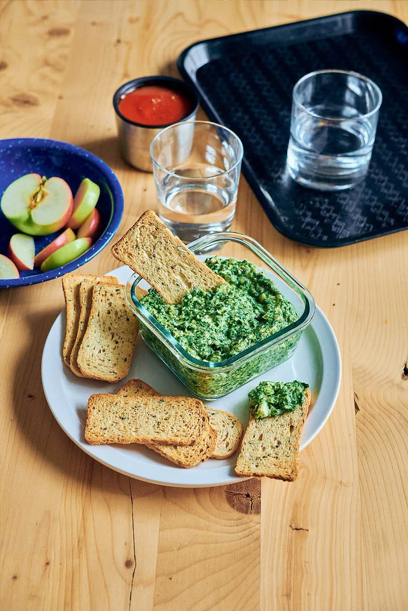 Boulangerie Grissol Melba Toast Sourdough Flavor with a spinach on a white plate with apples and water on a wooden table