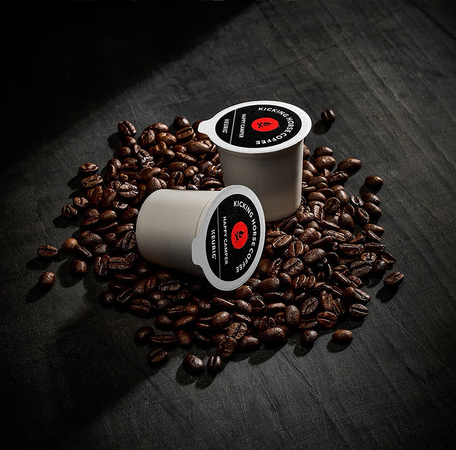 Two Kicking Horse Coffee single-serve cups surrounded by coffee beans on a dark surface