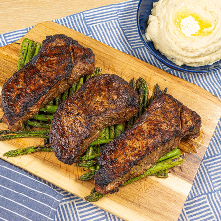Grilled steaks seasoned with Kinder's Buttery Steakhouse dry rub on a wood cutting board with asparagus and mashed potatoes