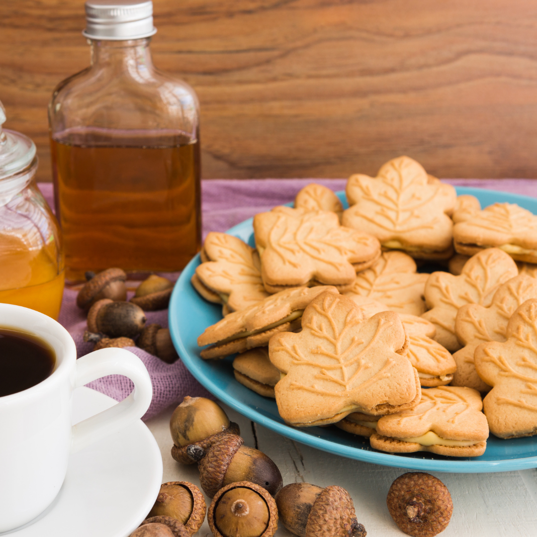 Maple leaf cookies on a blue plate with a bottle of syrup, jar of honey, and cup of coffee on a wooden surface.