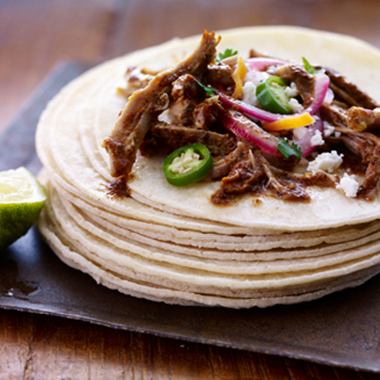Stack of tortillas with pulled meat, vegetables, and lime on a wooden surface