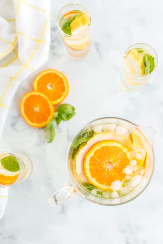 Glass pitcher and glasses of orange and lemon slices with ice on a marble surface.
