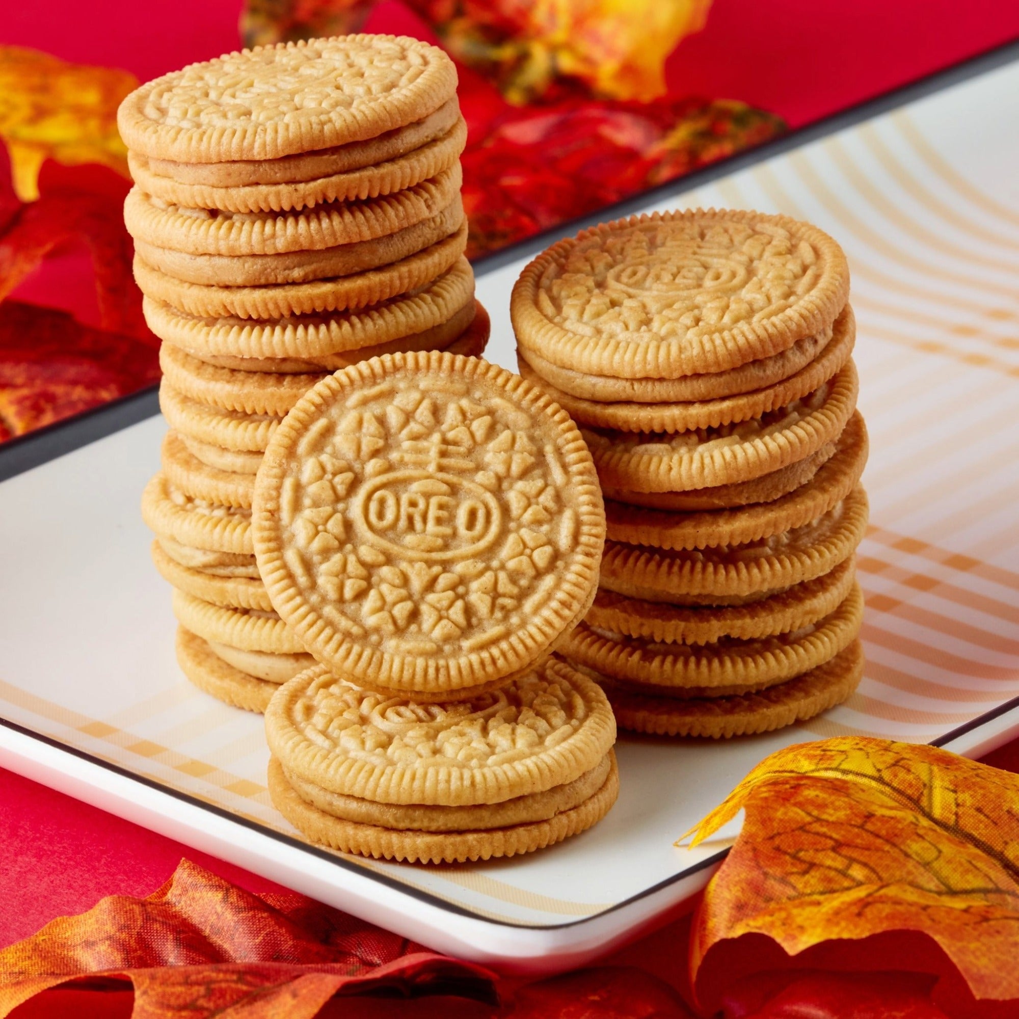 Three stacks of Oreo cookies with a single cookie in front, arranged on a white plate with autumn leaves and a red background.