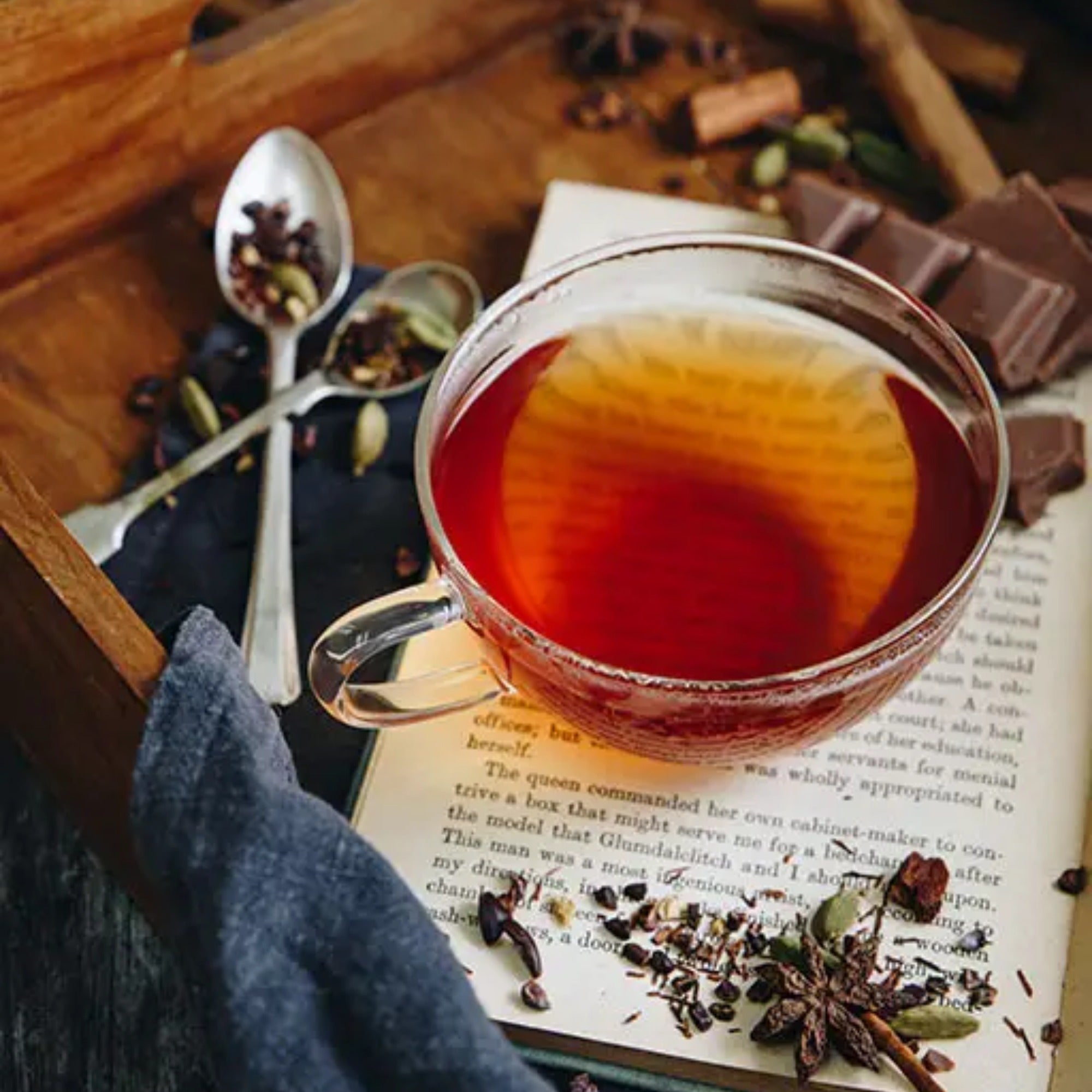 A cup of maple rooibos tea on an open book surrounded by pieces of chocolate, two tea spoons and herbs on a wooden tray.