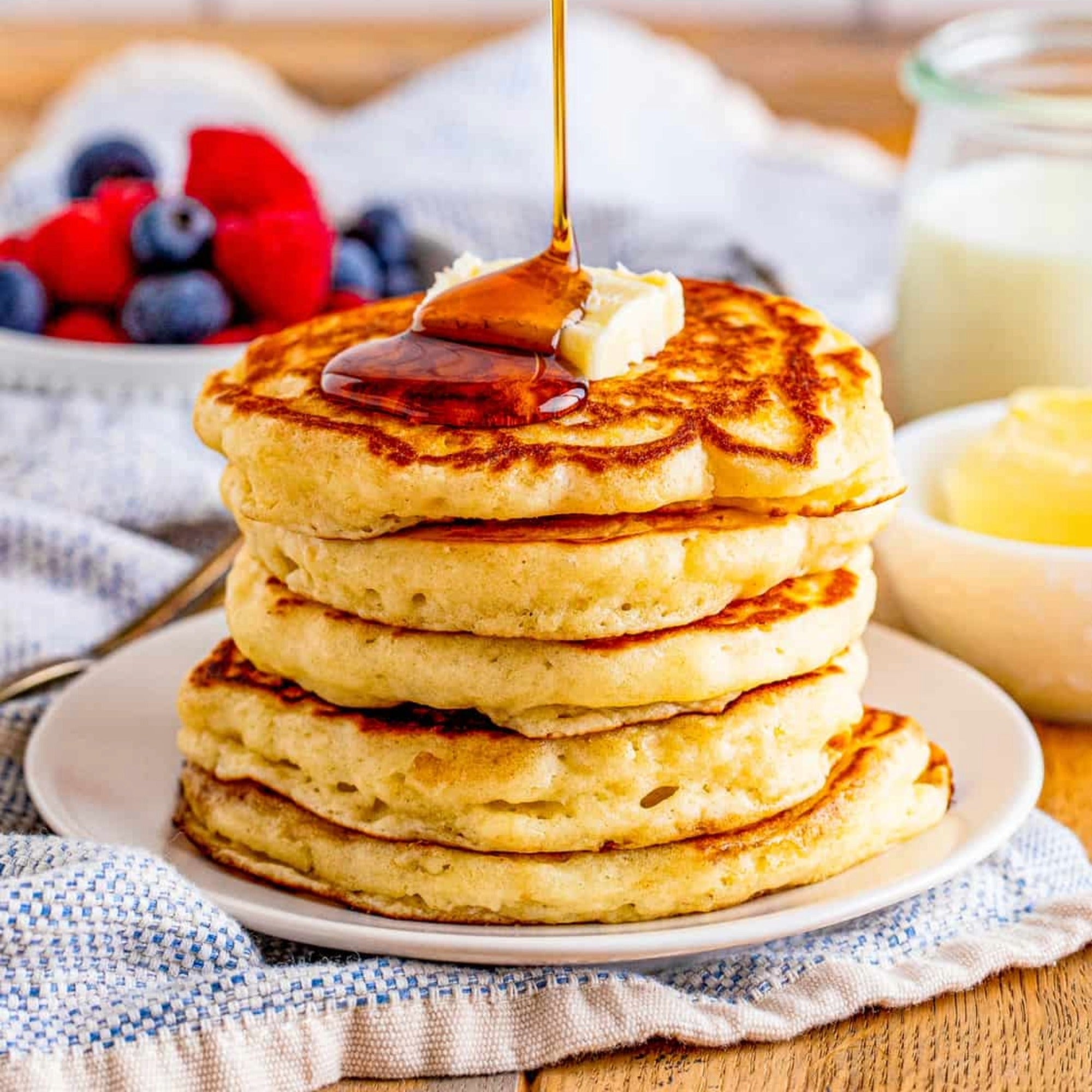 Stack of golden pancakes with butter and pouring syrup, with berries alongside.