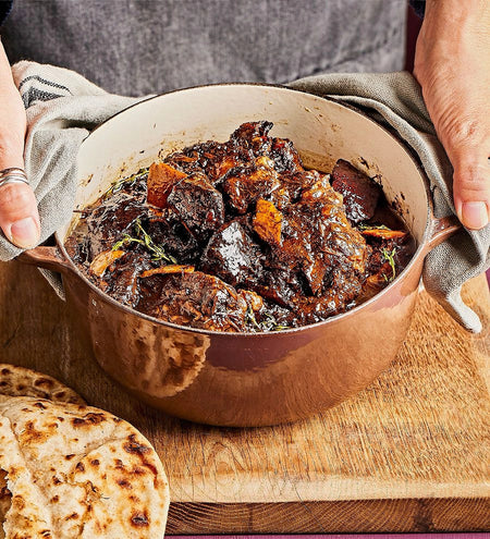 Hands holding a cooked Pepperpot stew in a cooking pot on a wooden surface with flatbread on the side