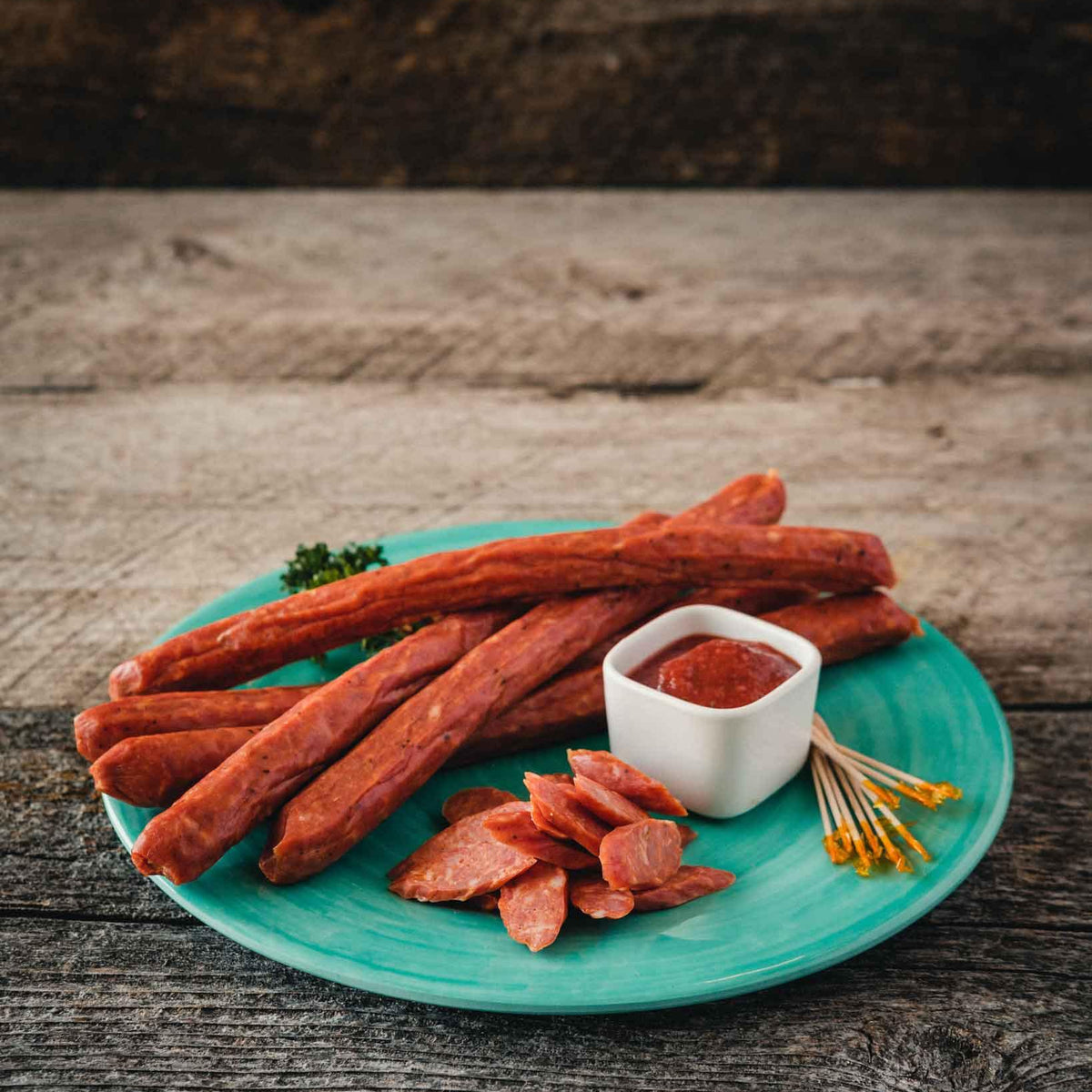 Pepperoni sausage sticks and tomato dip arranged on a turquoise plate with a wooden background 