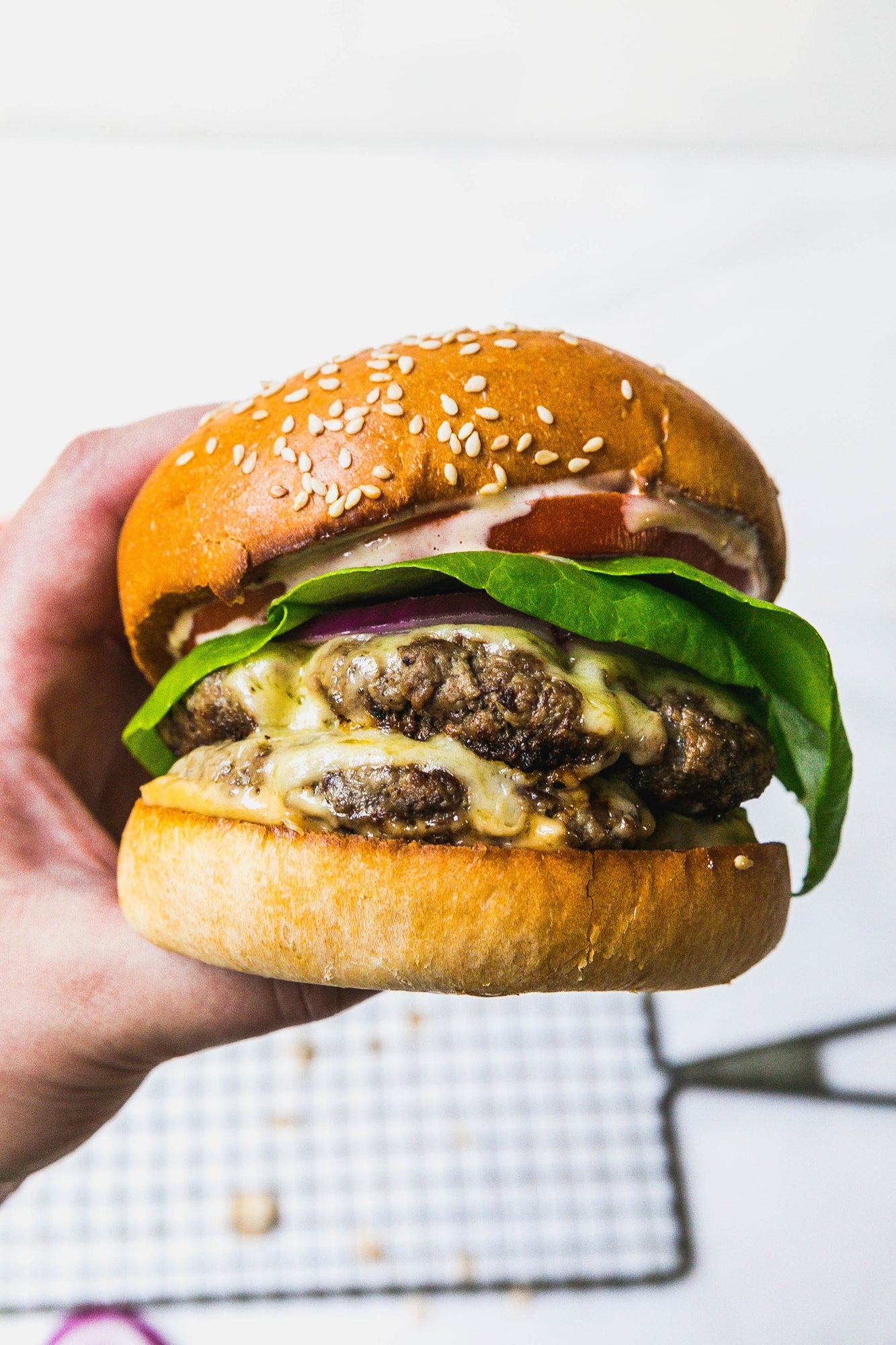 Hand holding a juicy burger with sesame seed bun, lettuce, cheese, and old fashioned dijon mustard on a white background