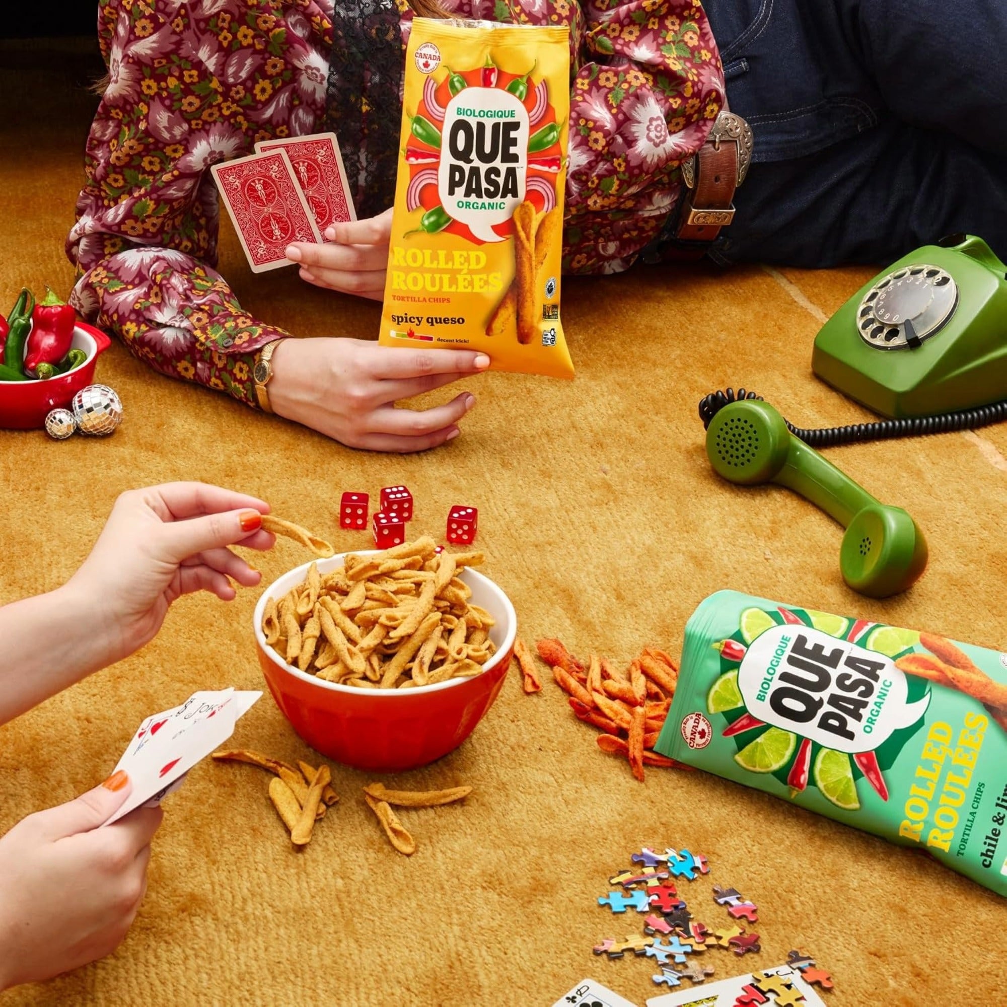 Que Pasa Spicy Queso and Chile & Lime rolled chips with a snack bowl, playing cards, dice, and puzzle pieces.