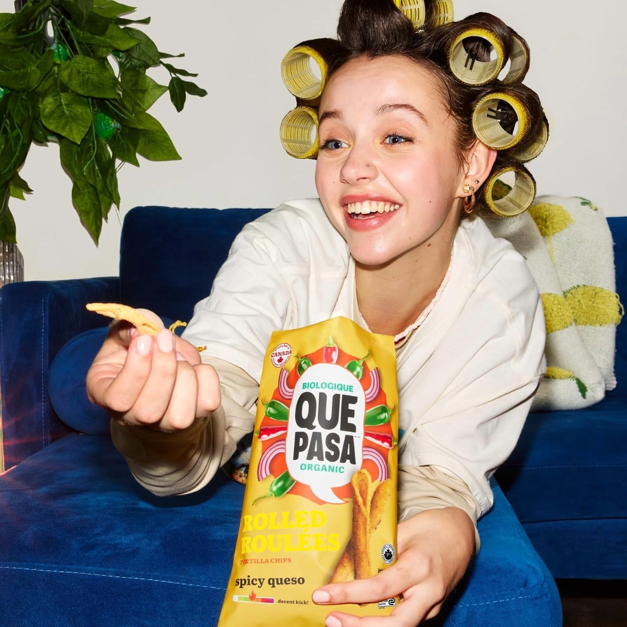 Person wearing hair rollers on a blue couch, holding a chip and a bag of Que Pasa Spicy Queso rolled tortilla chips.
