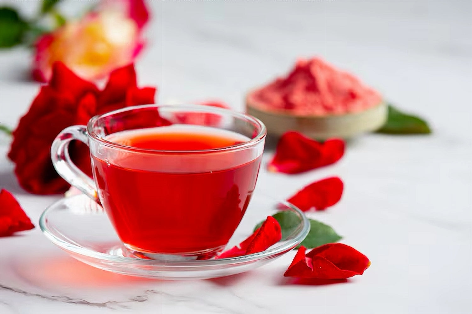 Clear glass cup filled with red rose orange pekoe tea, surrounded by red flowers and petals on a light background