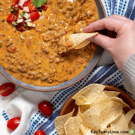 Hand dipping a tortilla chip into a bowl of queso fundido with tomatoes and cilantro on a blue and white checkered tablecloth.