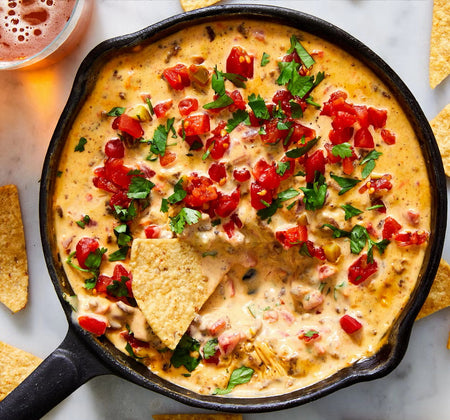 Hot cheese dip with tomatoes and cilantro in a skillet with a tortilla chip dipped into it.