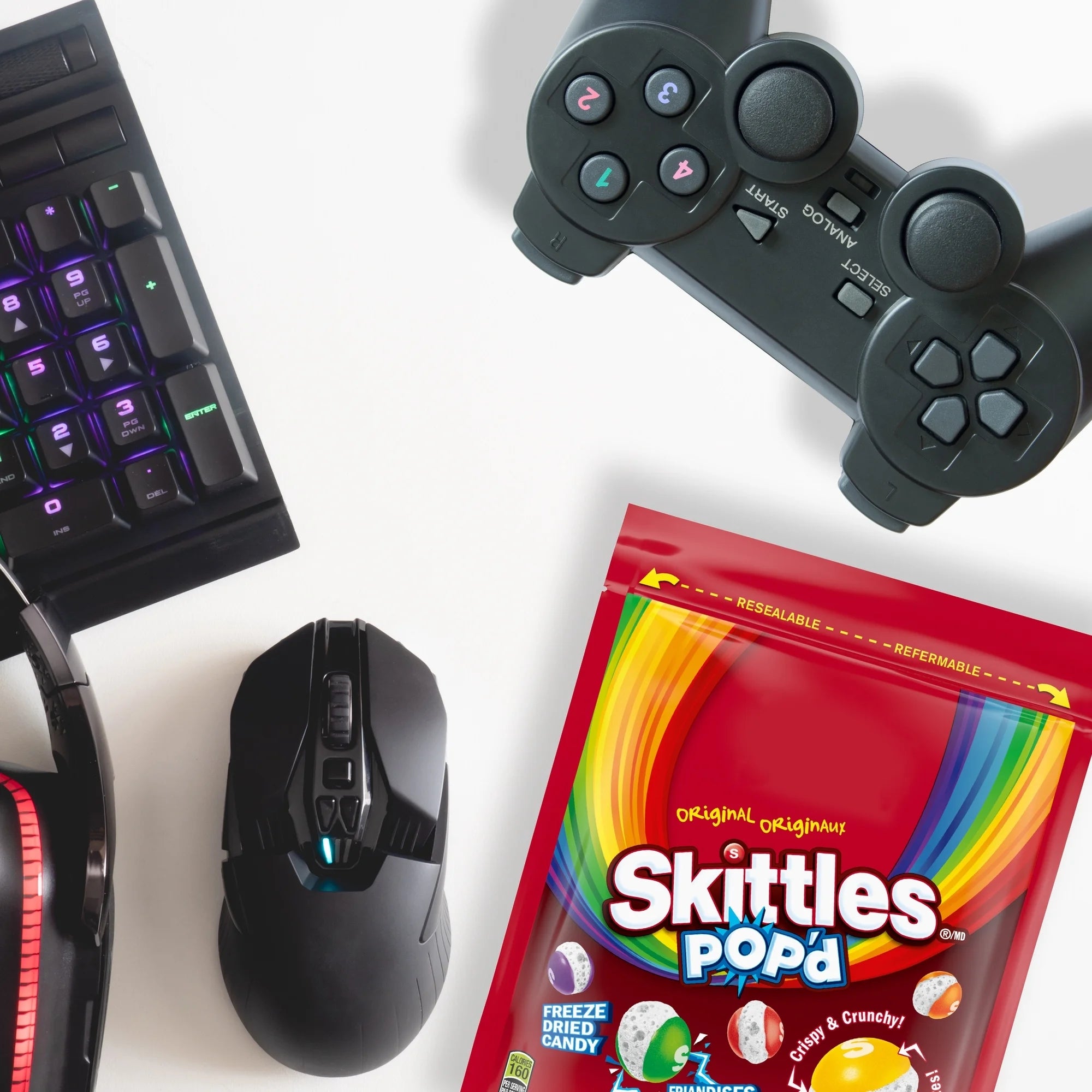 Gaming setup with keyboard, mouse, and controller on a white background with Skittles Pop-a package.