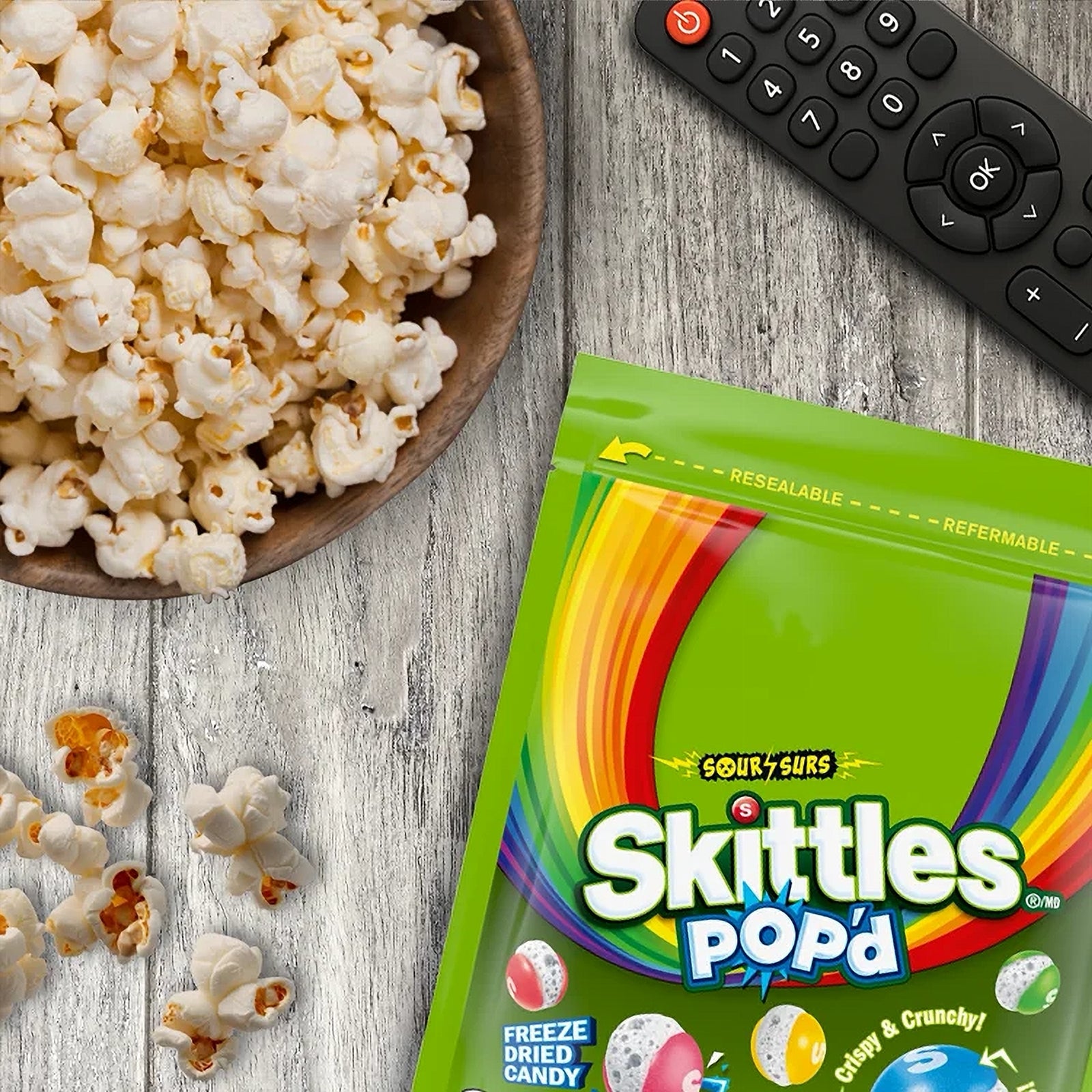 Skittles Pop'd candy package with a bowl of popcorn and remote control on a wooden surface