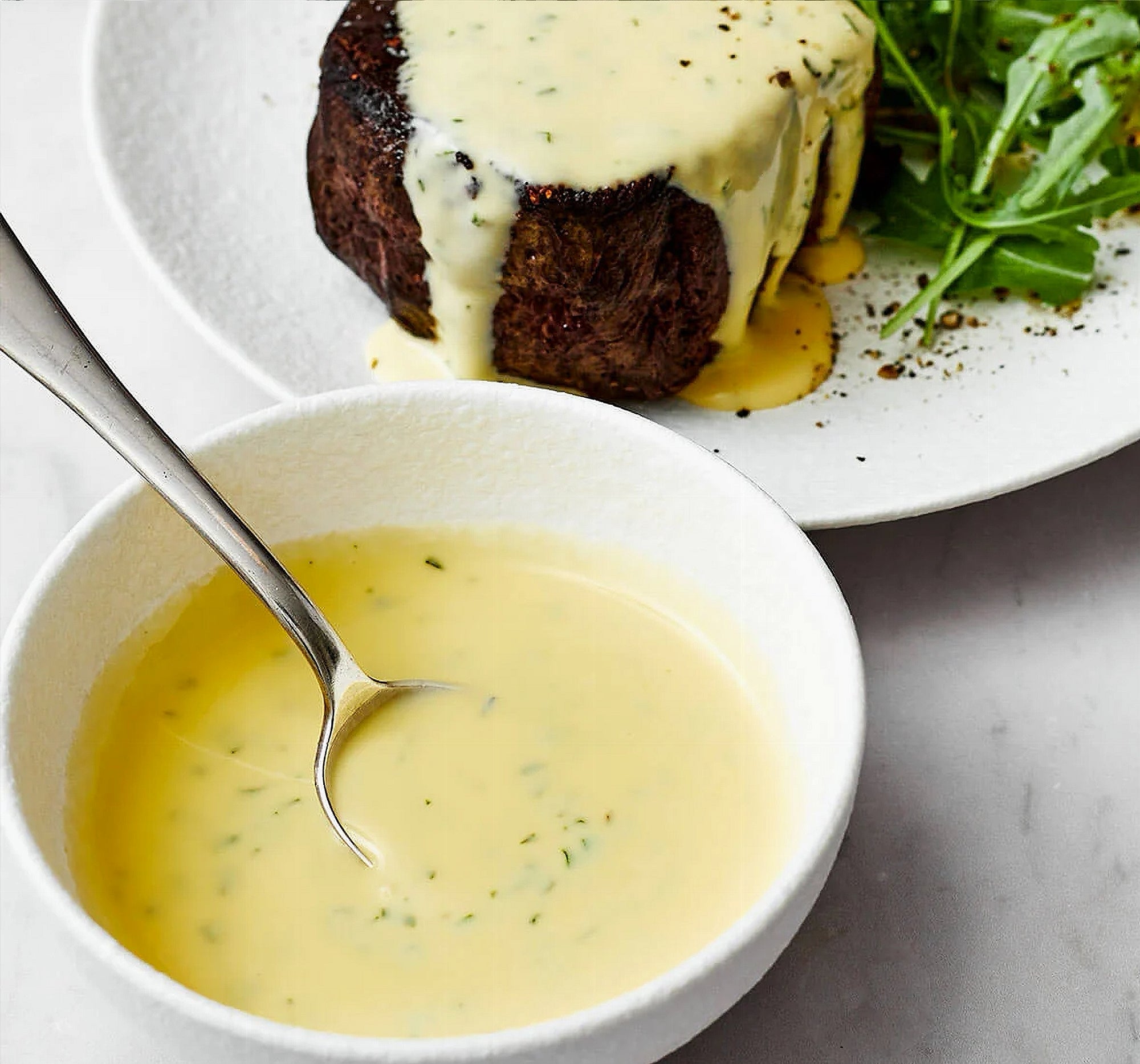 Bowl of The Keg Bearnaise Sauce sauce with a spoon, accompanied by a plated dish with a steak and arugula.