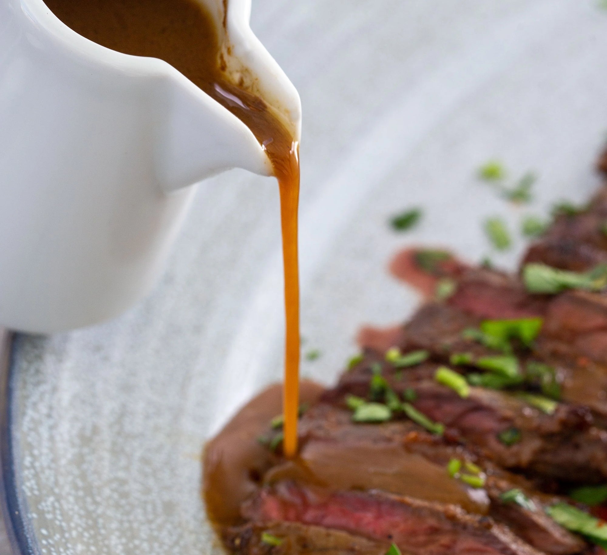 Demi glace gravy being poured from a white pitcher onto a plate of steak with green herbs.