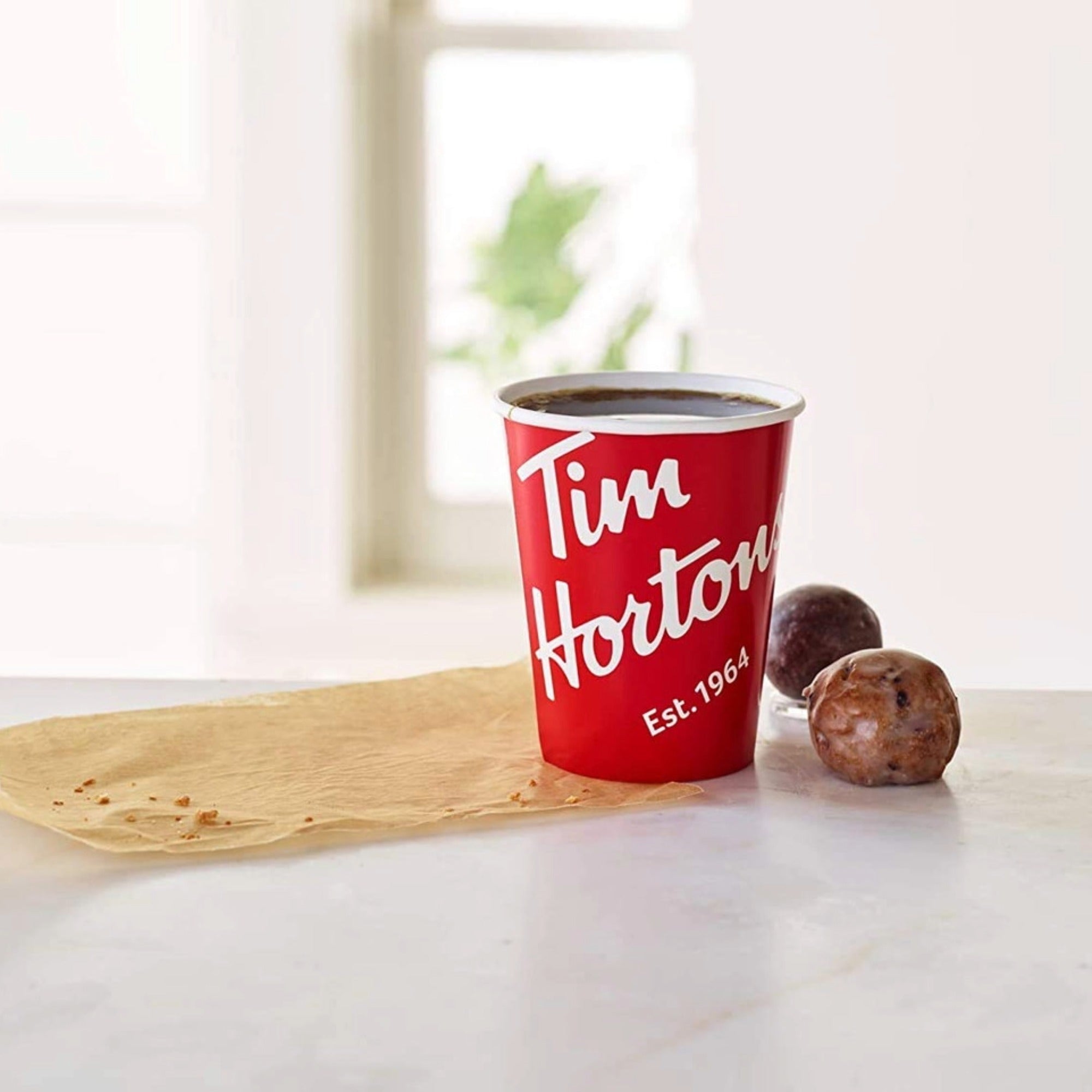 A red Tim Hortons cup filled with Tim Hortons maple coffee, two Timbits, standing on a piece of parchment paper, on a white surface.