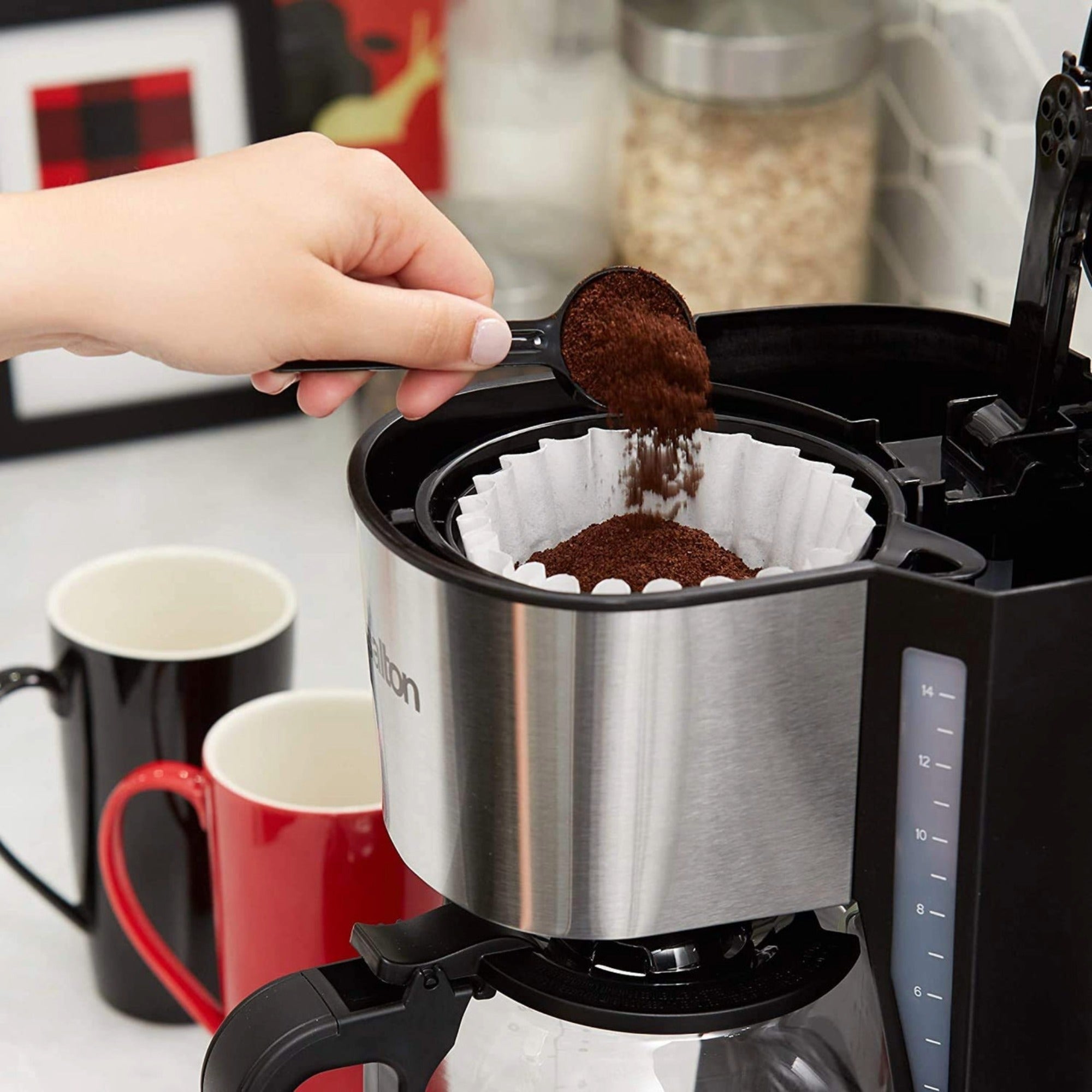A person who fills ground coffee into a coffee maker, one red and one black mug standing beside the coffee maker on a white counter in the kitchen.
