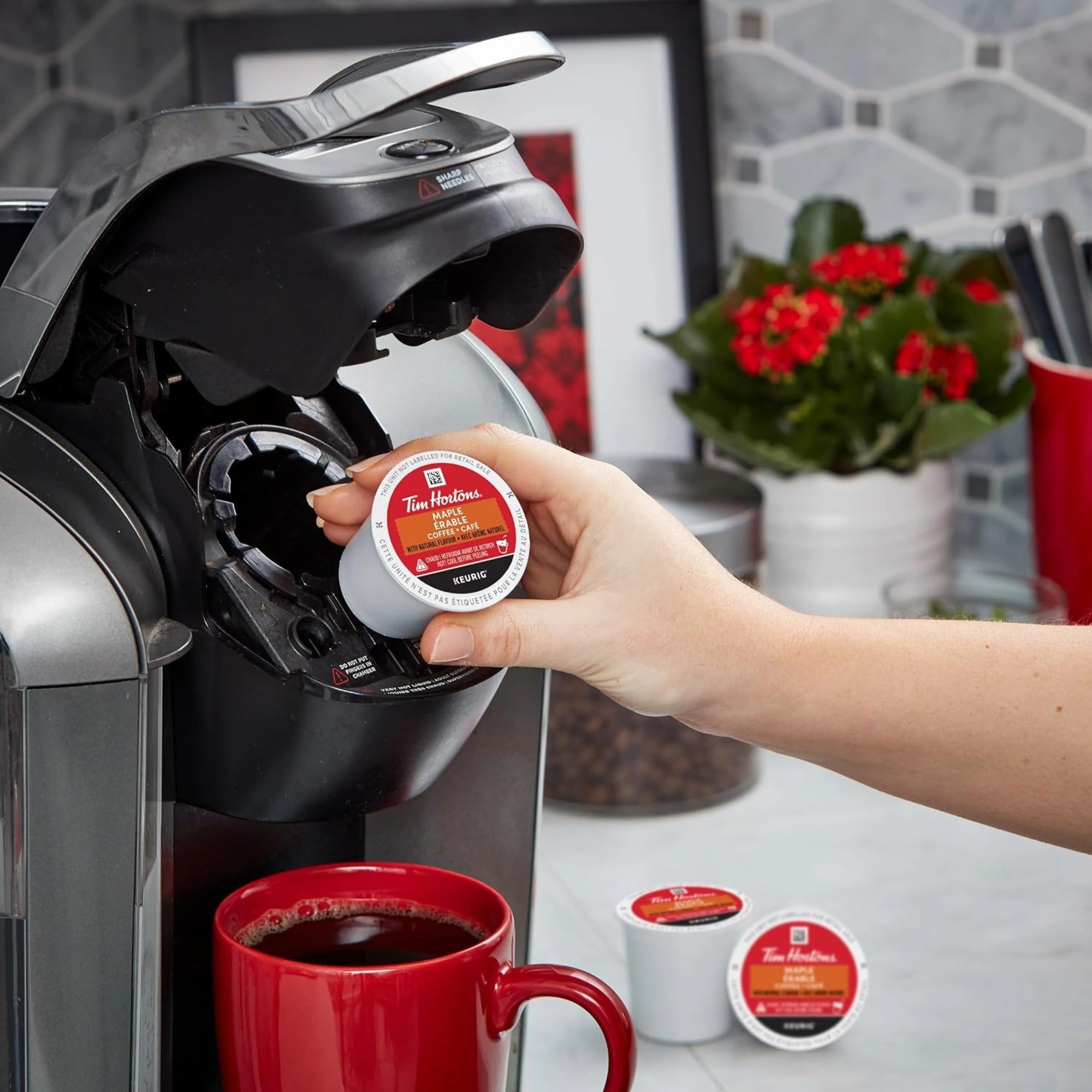 Person inserting a coffee pod into a Keurig machine with a red mug filled with coffee on a kitchen counter.