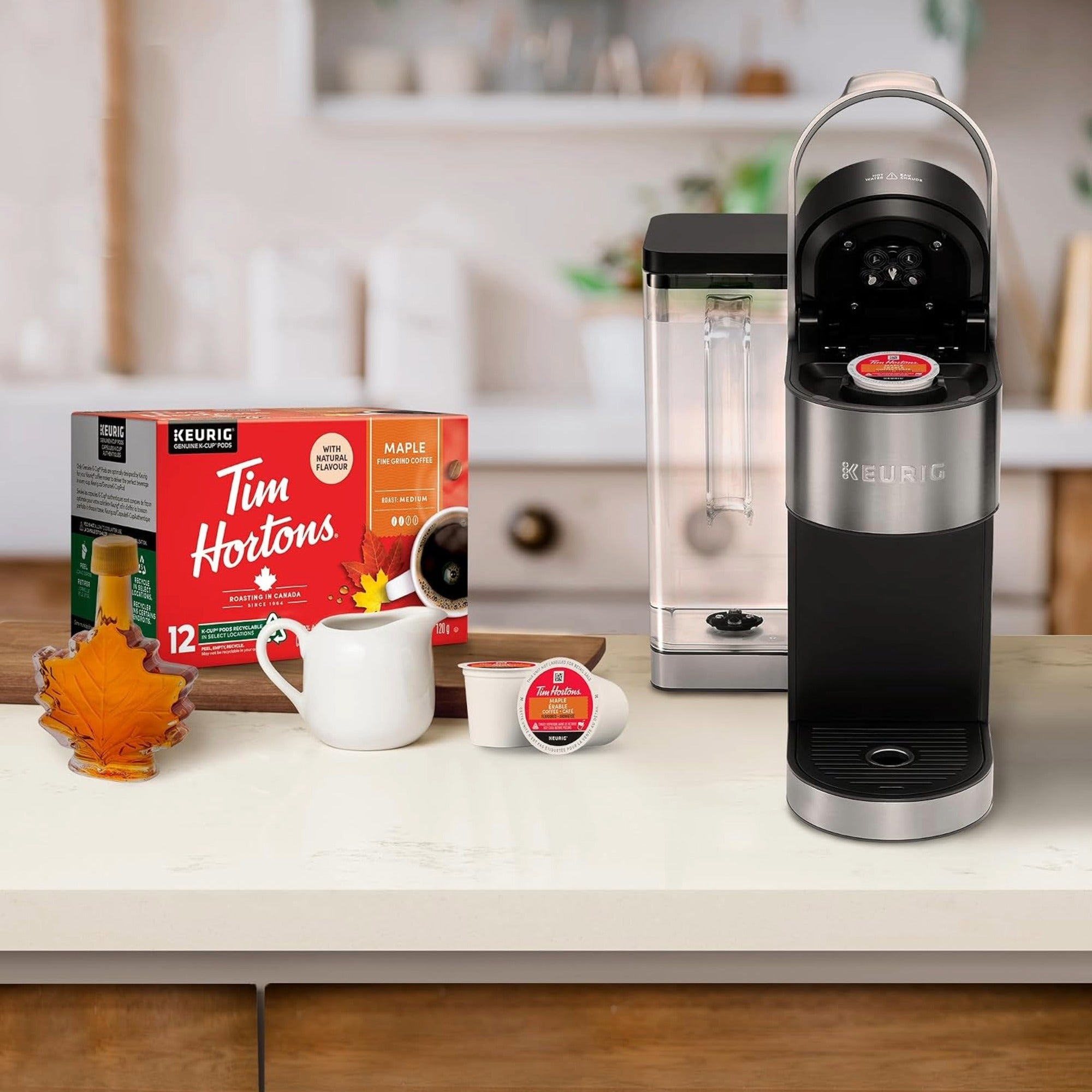 Keurig coffee machine on a kitchen counter next to a Keurig Tim Hortons coffee pod box, a white mug, a coffee pod, and a maple leaf-shaped glass on a light countertop.