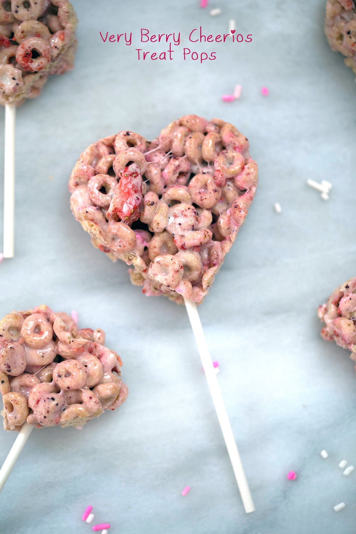 Heart-shaped Very Berry Cheerios treats on sticks with pink and white sprinkles on a light blue background.