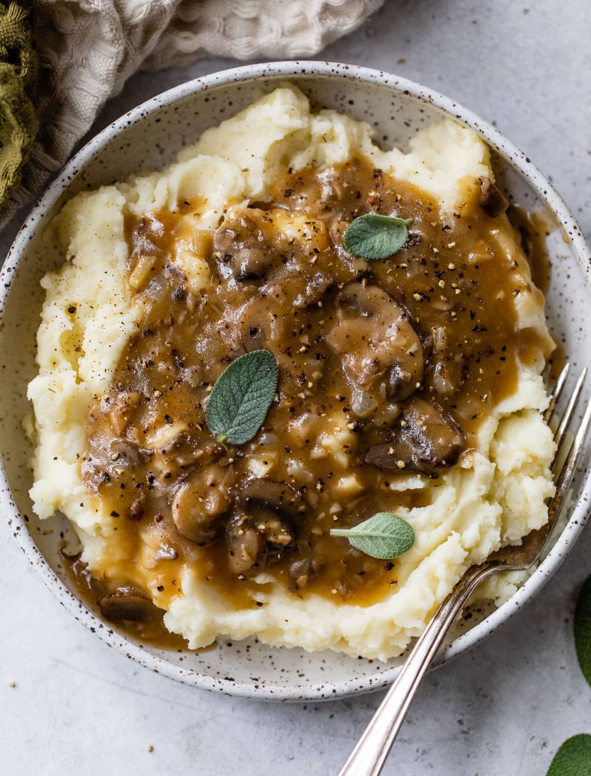 Mashed potatoes with brown gravy and fresh herbs in a speckled bowl on a light background