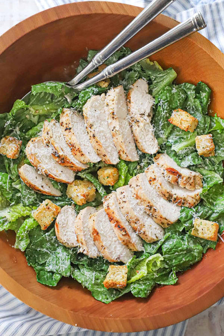 Caesar salad topped with chicken meat and two silver spoons in a wooden bowl on a white and blue striped cloth