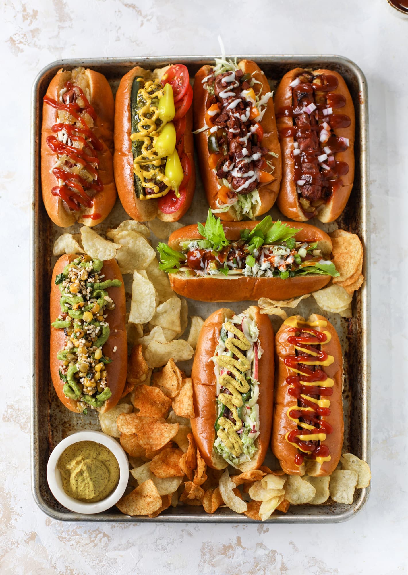 Assorted hot dogs with various toppings on a baking tray with chips and condiments.
