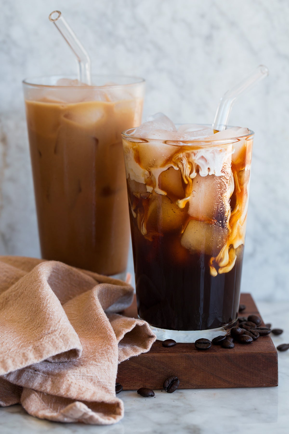 Two glasses of iced coffee with straws on a marble surface with a beige towel and coffee beans.